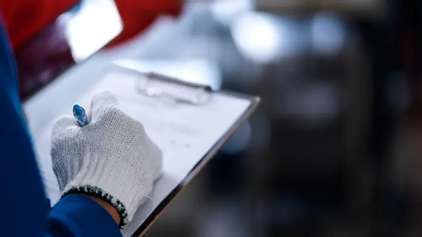 Inspector con guantes de seguridad escribiendo en un portapapeles durante una auditoría en la industria agroalimentaria.