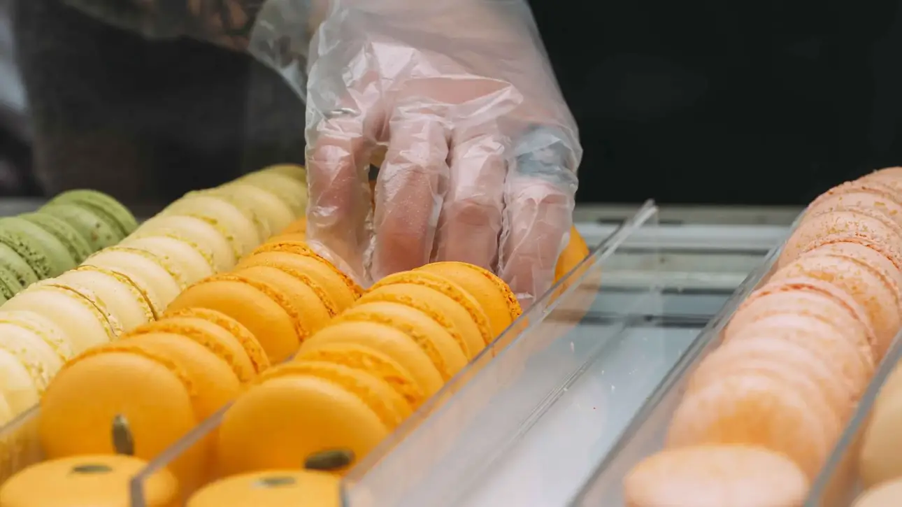 Mano con guantes de plástico transparente manipulando macarons en vitrina de pastelería