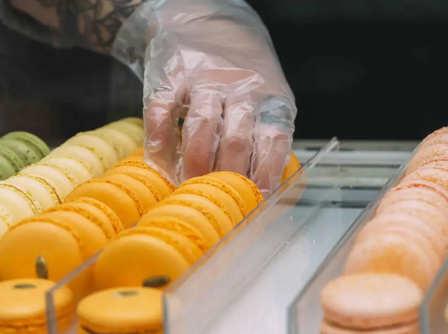 Mano con guantes de plástico transparente manipulando macarons en vitrina de pastelería